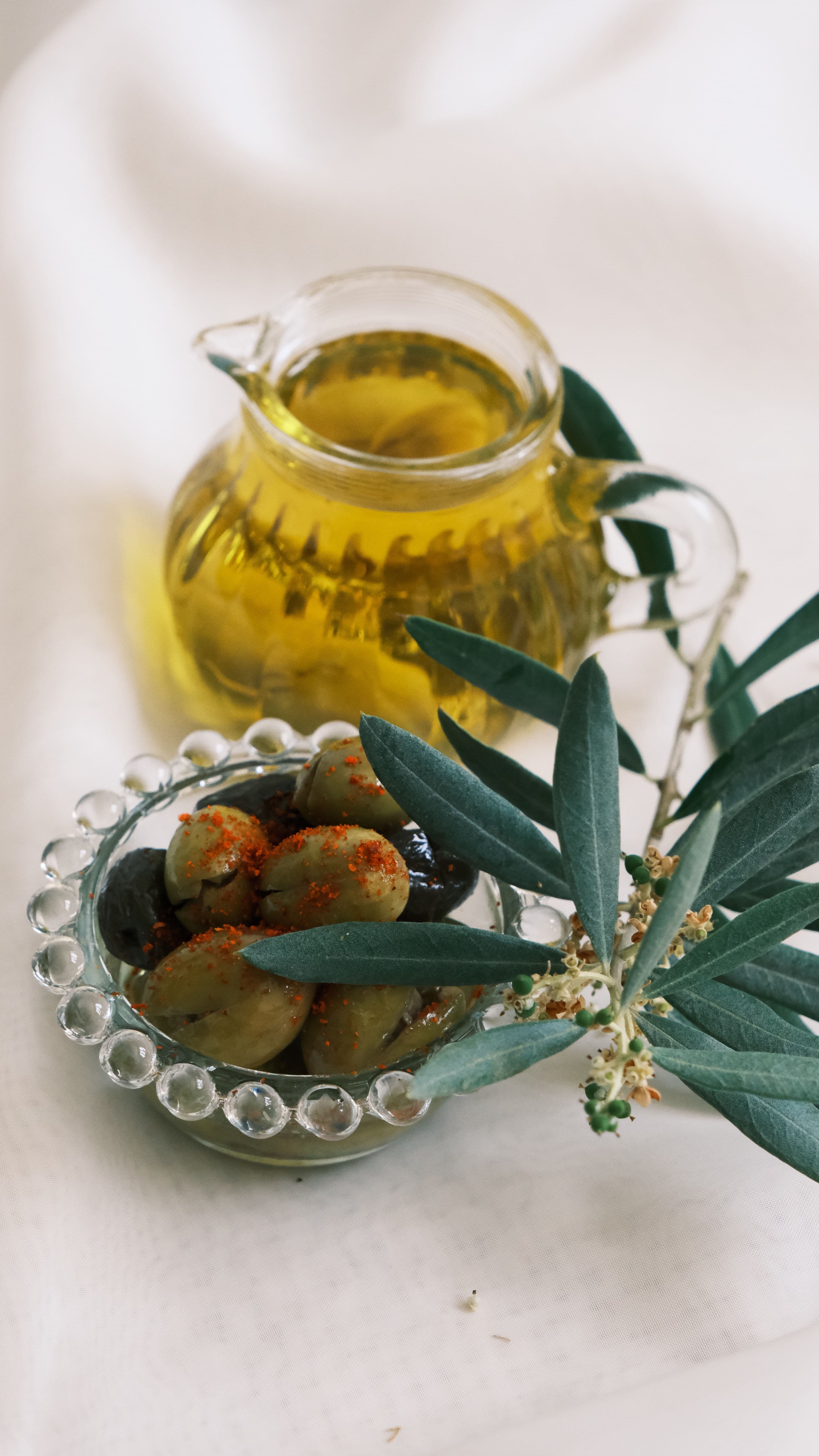 Cam sürahide altın renkli natürel sızma zeytinyağı, yanında zeytin dalı ve pul biberli kırma yeşil zeytin